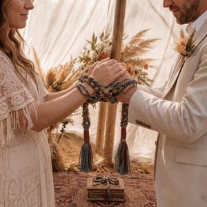 Könnte beinhalten: Ein Handfasting-Seil aus geflochtenem, mehrfarbigem Garn in Blau-, Grau- und Beigetönen. Das Seil hat an den Enden zwei dicke Quasten, die mit dunkelrotem Garn umwickelt sind, und ist um zwei zusammengehaltene Hände geschlungen.