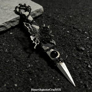 May include: A silver dagger with a pointed blade, black accents, and a decorative handle. The handle features a black gem, black roses, and a butterfly-like design. A chain connects to the top of the dagger.