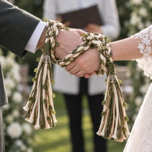 Könnte beinhalten: Ein Handfasting-Seil aus geflochtenem cremefarbenem und olivgrünem Seil, verziert mit kleinen grünen Stoffblättern. Jedes Ende ist mit einem silberfarbenen Lebensbaum-Anhänger und einer dicken Quaste aus cremefarbenen und grünen Seilsträngen versehen.