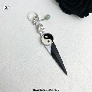 May include: A black and white pendant with a yin-yang symbol, coins, and a green bead. The pendant has a sharp, blade-like design and a white ring at the top. The text "D6" is in the upper left corner.
