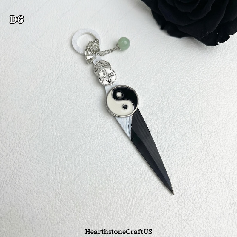 May include: A black and white pendant with a yin-yang symbol, coins, and a green bead. The pendant has a sharp, blade-like design and a white ring at the top. The text "D6" is in the upper left corner.
