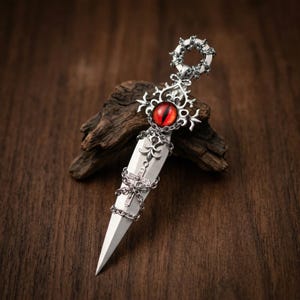 May include: A silver dagger with a red eye-shaped gem in the center. The dagger features intricate decorative elements, including a chain wrapped around the blade and a circular handle. The dagger is displayed on a wooden surface.