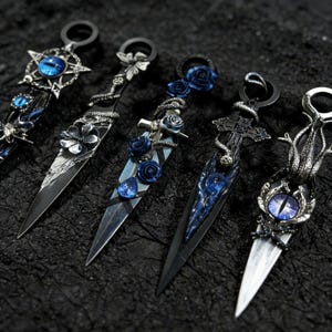May include: Close-up of five decorative daggers with intricate designs. Each dagger features a silver blade and a black handle with unique embellishments, including blue gemstones, flowers, and serpent motifs. The daggers are displayed on a textured black surface.