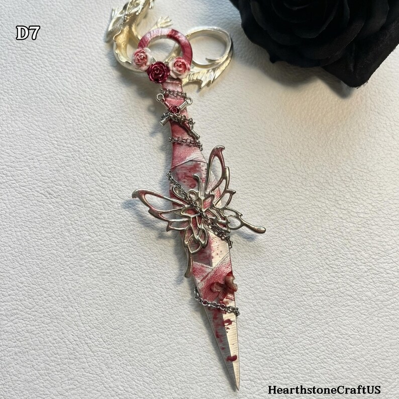 May include: A decorative dagger with a silver blade and a pink and white handle. The handle is adorned with small red roses and a silver butterfly. A silver chain wraps around the handle. The text "D7" is in the upper left corner.