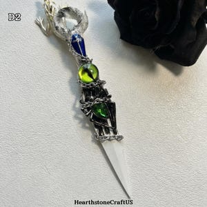 May include: A decorative dagger with a white blade and a detailed handle. The handle features a silver-toned skeleton hand design, adorned with green and yellow gemstones, and a blue gem. The top of the dagger has a silver-toned lizard design. The text "HearthstoneCraftUS" is at the bottom.