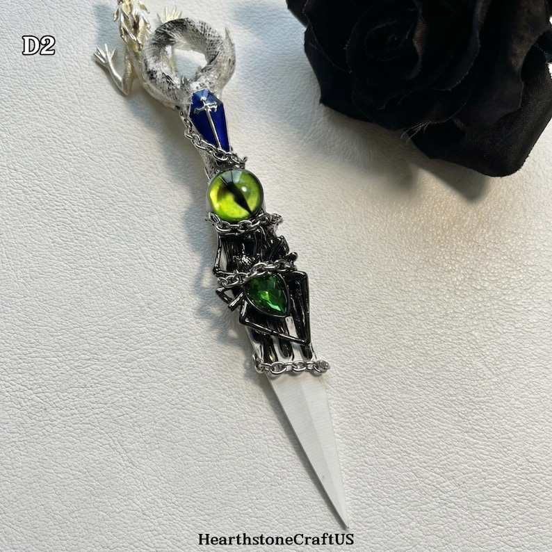 May include: A decorative dagger with a white blade and a detailed handle. The handle features a silver-toned skeleton hand design, adorned with green and yellow gemstones, and a blue gem. The top of the dagger has a silver-toned lizard design. The text "HearthstoneCraftUS" is at the bottom.
