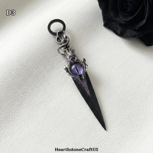 May include: A decorative dagger pendant with a black blade and a purple eye-shaped gem. The handle features a snake and cross design. The pendant is on a white surface. The text "HearthstoneCraftUS" is at the bottom left.