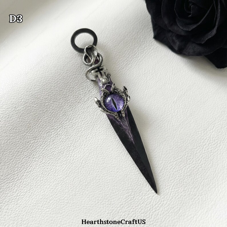 May include: A decorative dagger pendant with a black blade and a purple eye-shaped gem. The handle features a snake and cross design. The pendant is on a white surface. The text "HearthstoneCraftUS" is at the bottom left.