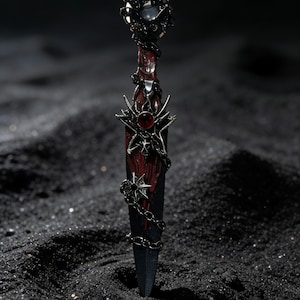 May include: A decorative dagger with a black blade and a red handle. The handle is adorned with silver-colored embellishments, a red gem, and a chain. The dagger's pommel features a dark, ornate design. The dagger is set against a black background.
