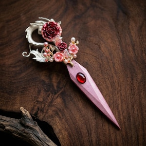 Könnte beinhalten: Eine dekorative Haarnadel mit einem silbernen Drachendesign und einem rosa Holzgriff. Die Nadel hat eine rote Rose, rosa Blüten, Perlen und einen roten Edelstein. Das Haaraccessoire ist vor einem dunklen Holzhintergrund platziert.