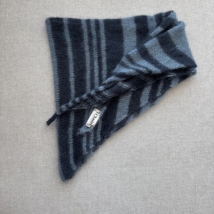 Puede incluir: Una bufanda de punto a rayas azules y grises, doblada en forma de triángulo. La bufanda tiene una etiqueta que dice "I.C. knit's". La bufanda está hecha de un hilo suave y texturizado.