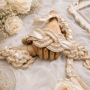 May include: A decorative braided rope belt or sash featuring interwoven beige and white cord with integrated cream-coloured satin ribbon accents. The braided textile accessory is draped over a wooden mannequin hand, showcasing a complex knot design.