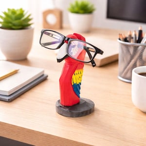 May include: A wooden parrot eyeglass holder, hand-carved and painted in bright colours. The parrot is predominantly red with yellow and blue wing details. The holder, with black-framed glasses, sits on a dark grey base. A decorative desk accessory.