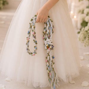 Pode incluir: Um acessório de cordão branco trançado com pequenas flores de tecido coloridas em tons de rosa, azul e amarelo, acentuado com pequenas folhas verdes. O cordão é atado no centro e pende em várias tiras adornadas com detalhes florais.
