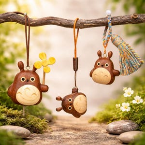 May include: Three wooden Totoro-inspired ornaments hanging from a branch. The ornaments are brown with light wood bellies and faces. One holds a yellow flower, another has a tassel, and the third is upside down.