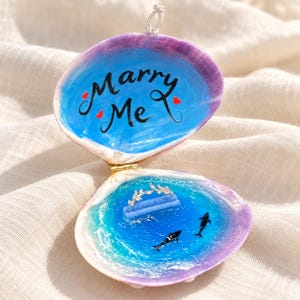 May include: An open seashell ring box with a purple and white exterior. The interior features a blue ocean painting with the text Marry Me and two small red hearts. A blue velvet ring holder and two miniature orca figures are inside.