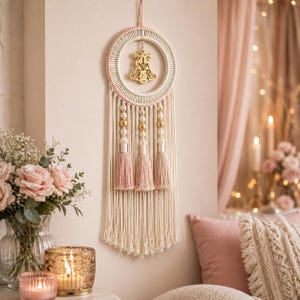 May include: A macrame wall hanging featuring a circular frame wrapped in cream cord, a gold-toned bell ornament suspended in the centre, and three hanging strands adorned with gold beads and dusty pink tassels, finished with long cream fringe.