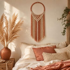 Peut inclure: Une décoration murale circulaire composée de cordes en macramé alternant des bandes verticales crème et terre cuite. Le motif présente un nœud en forme de V sur le haut, avec de longues franges tombant d'un cadre en cerceau de bois.