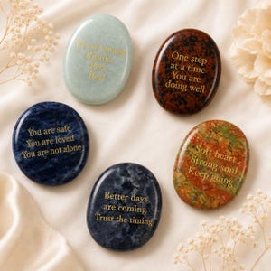 Pode incluir: Cinco pedras polidas de formato oval em várias cores, incluindo verde-claro, castanho-escuro, azul-profundo e verde-avermelhado manchado. Cada pedra apresenta texto gravado em dourado com frases motivacionais como You are enough, One step at a time, You are safe, Soft heart e Better days are coming.