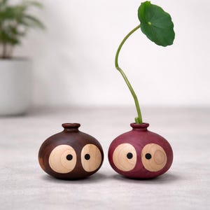 May include: Two small, round wooden vases with a smooth finish. One vase is dark brown, and the other is a deep purple. Both feature two large, light-coloured circular inlays on the front, each with a small black dot in the centre, resembling eyes.