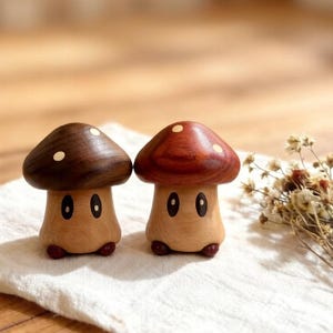 Puede incluir: Dos figuras de hongos de madera con sombreros marrones y rojizos, cada uno con puntos blancos. Los tallos de los hongos son de madera clara con ojos pintados y pequeños pies rojos. Sobre un paño blanco.