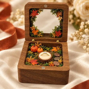 Può includere: Un portaanelli quadrato in legno con coperchio incernierato e finestra in vetro. L'interno è decorato con muschio verde, zucche arancioni, piccoli sassi e foglie autunnali. Un anello dorato con una pietra chiara poggia su un cuscinetto beige.
