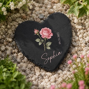 Peut inclure: Une plaque en ardoise noire en forme de cœur ornée d'une illustration imprimée d'une rose rose avec des feuilles vertes. Le prénom Sophia est écrit en écriture cursive blanche et la date 22.04.2021 est imprimée verticalement en police blanche.