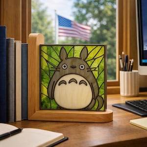 May include: A wooden bookend featuring a stained glass panel with a cartoon character design. The character is gray with a white belly and large eyes, set against a green leaf background. The bookend is on a wooden desk with books and a notepad.