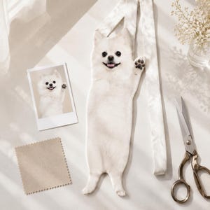 May include: A long, narrow white fabric pouch featuring a realistic photographic print of a fluffy white Pomeranian dog with its paw raised. The pouch includes a matching white fabric strap, a small square fabric swatch, and a small photo print.