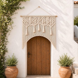May include: A cream-colored macrame wall hanging suspended from a wooden dowel, featuring intricate knotted patterns, diamond shapes, and hanging tassels. The textile art is composed of long, vertical cotton cords that form an arched shape.