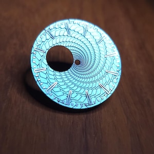 K&ouml;nnte beinhalten: Ein runder, t&uuml;rkisfarbener Zifferblatt mit einem Spiralmuster und silberfarbenen Markierungen. Das Zifferblatt hat ein zentrales Loch und liegt auf einer dunklen Holzoberfl&auml;che.