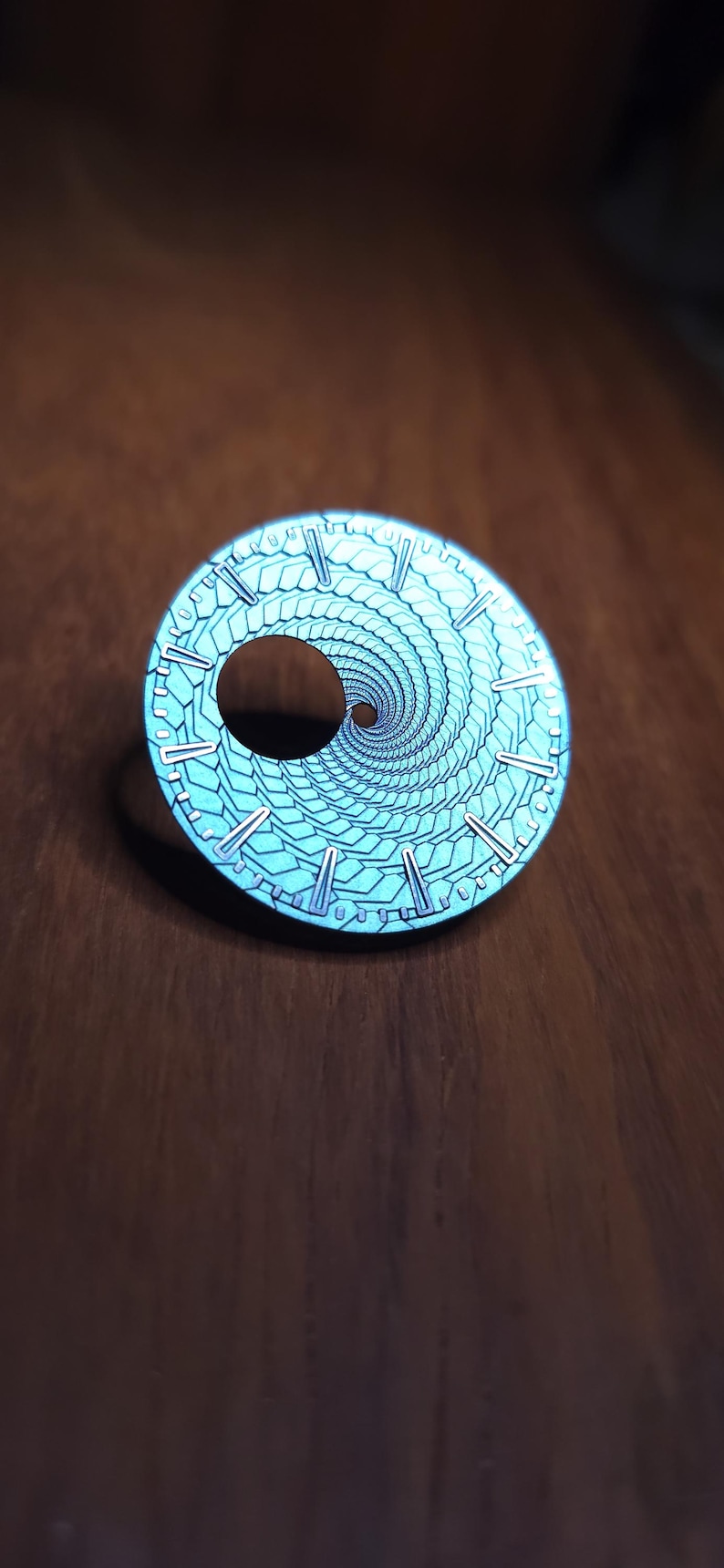 K&ouml;nnte beinhalten: Ein runder, t&uuml;rkisfarbener Zifferblatt mit einem Spiralmuster und silberfarbenen Markierungen. Das Zifferblatt hat ein zentrales Loch und liegt auf einer dunklen Holzoberfl&auml;che.