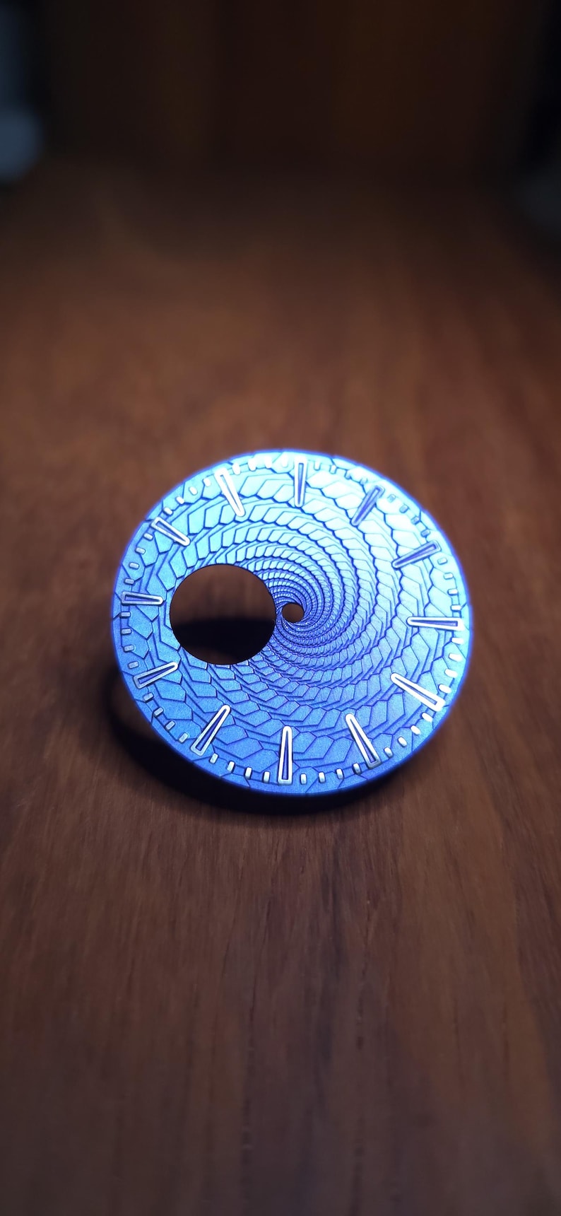 Op de afbeelding: Een blauwe wijzerplaat met een spiraalvormig ontwerp en een centrale opening. De wijzerplaat heeft een textuurpatroon en witte markeringen langs de rand. De wijzerplaat ligt op een houten oppervlak.