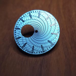 K&ouml;nnte beinhalten: Ein silberfarbenes Zifferblatt mit einem einzigartigen Spiralmuster. Das Zifferblatt hat eine runde Form mit einem zentralen Loch und ist mit kleinen Linien f&uuml;r die Stunden markiert. Das Spiralmuster erzeugt einen faszinierenden Effekt.
