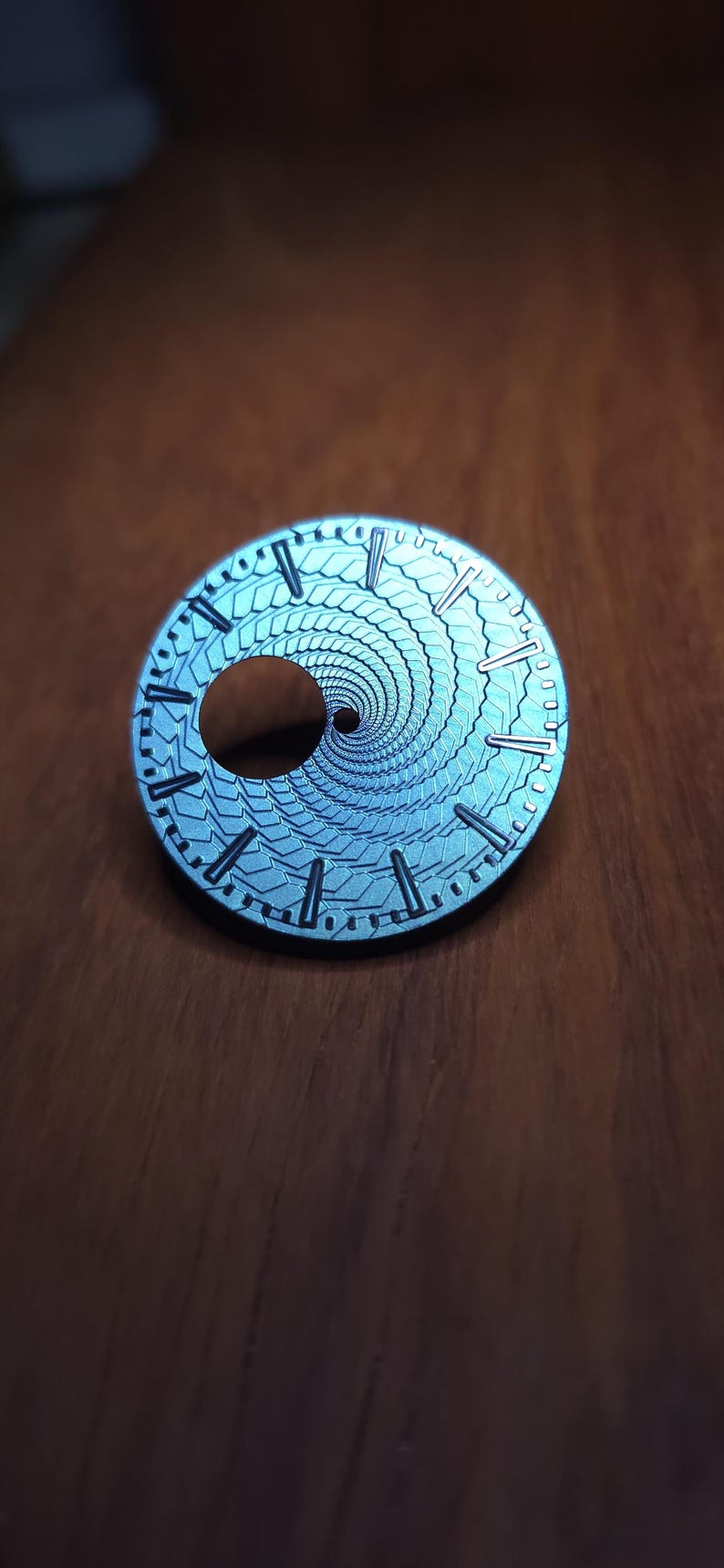 K&ouml;nnte beinhalten: Ein silberfarbenes Zifferblatt mit einem einzigartigen Spiralmuster. Das Zifferblatt hat eine runde Form mit einem zentralen Loch und ist mit kleinen Linien f&uuml;r die Stunden markiert. Das Spiralmuster erzeugt einen faszinierenden Effekt.
