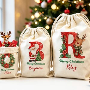 Pode incluir: Três sacos de presente de lona em tom off-white com cordão de tamanhos variados. O menor apresenta uma guirlanda e o nome Olivia. O saco médio mostra uma letra B, o Papai Noel e o nome Benjamin. O maior exibe uma letra R, uma rena e o nome Riley. Todos os sacos incluem o texto Merry Christmas.