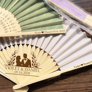 May include: Several decorative hand fans in various colors, including white, green, and purple. Some fans feature the names "Violet & Daniel" and the date "09.16.2028". One fan has a silhouette of a couple and floral accents.