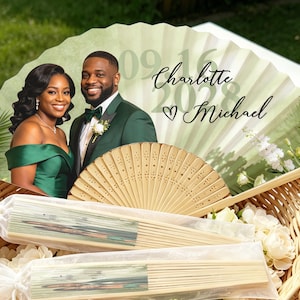 May include: A folding hand fan featuring a printed photograph of a couple in formal green attire, with the text Charlotte and Michael, a heart icon, and the date 09.16.2028. The fan has a light wood frame and is displayed with two additional fans.