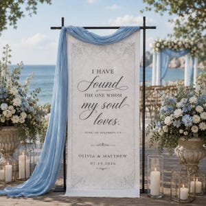 May include: A vertical rectangular wedding sign featuring the text I HAVE FOUND THE ONE WHOM MY SOUL LOVES, SONG OF SOLOMON 3:4, OLIVIA & MATTHEW, and 07.19.2026. The sign is draped with light blue fabric on a black metal stand.