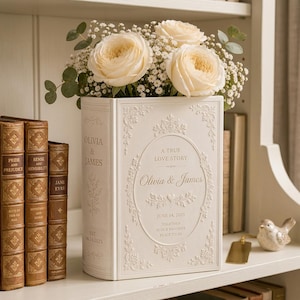 May include: A cream-colored book-shaped vase featuring embossed floral borders and the text A TRUE LOVE STORY, Olivia & James, JUNE 14, 2025, TOGETHER IS OUR FAVORITE PLACE TO BE, filled with white roses and baby's breath flowers.