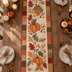 May include: A rectangular quilted table runner featuring a central cream-coloured panel decorated with embroidered orange, white, and green pumpkins, autumn leaves, and berries, bordered by a patchwork frame of various autumn-coloured fabric squares.