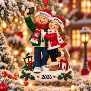 May include: A Christmas ornament featuring two figures wearing red Santa hats with the names Benjamin and Delaney printed on them. The figures are taking a selfie, wearing winter clothing, and standing on a snowy base with the year 2026 printed on it.