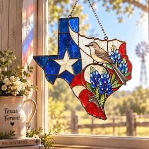 May include: A stained glass suncatcher shaped like the state of Texas, featuring a blue, white, and red flag design with a central white star. It depicts a perched mockingbird and bluebonnet flowers, suspended by a metal chain from a suction cup.