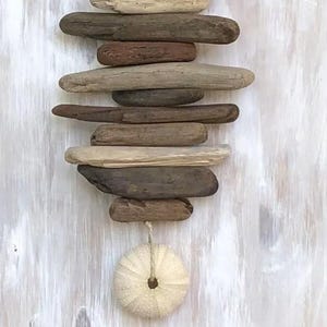 May include: A vertical wall hanging composed of multiple pieces of weathered driftwood in varying shades of brown and gray, arranged horizontally and stacked. A single, round, off-white sea urchin shell is suspended from the bottom piece by a string.