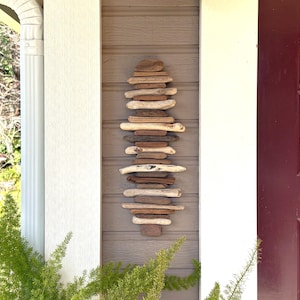 May include: A vertical wall art piece made of stacked driftwood pieces in shades of brown and beige. The artwork is mounted on a brown wooden wall, flanked by white columns and a red door.