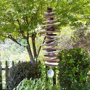 May include: A vertical hanging mobile composed of numerous weathered driftwood pieces of varying lengths and shades of brown, stacked horizontally on a central cord, finished with a single clear, faceted glass crystal pendant at the bottom.