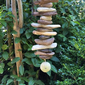 May include: A hanging driftwood mobile with a sea urchin at the bottom. The driftwood pieces are various shapes and shades of brown and tan. The mobile hangs in front of green foliage.