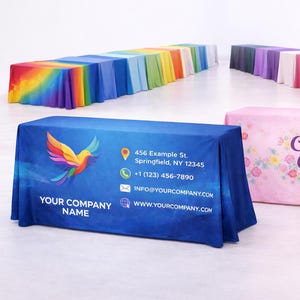 May include: Display table covers in various colors, including blue, pink, and rainbow. One cover features a colorful bird graphic and company information. Another displays the text "Cottage Crafts" with floral accents.