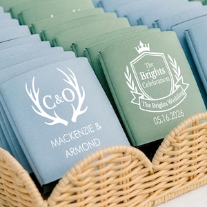 May include: A wicker tray holds stacks of blue and green wedding favors. Each favor has a white design and text, including the names "Mackenzie & Armond" and the date "05.16.2026". The tray sits on a white tablecloth.