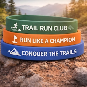 May include: Three stacked silicone wristbands in green, orange, and blue. Each band has white text and a graphic: "TRAIL RUN CLUB", "RUN LIKE A CHAMPION", and "CONQUER THE TRAILS". The bands are on a rocky surface.